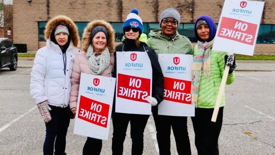 Five women standing together outside holding red and white placards that read on strike with the 十博官网在线 logo displayed on them in front of a the Magna building in Windsor, Ontario.