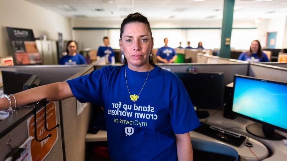 “一位女士站在她的办公室隔间里，穿着一件写着‘站起来捍卫你的王冠’的蓝色衬衫，同事们也这样做.”